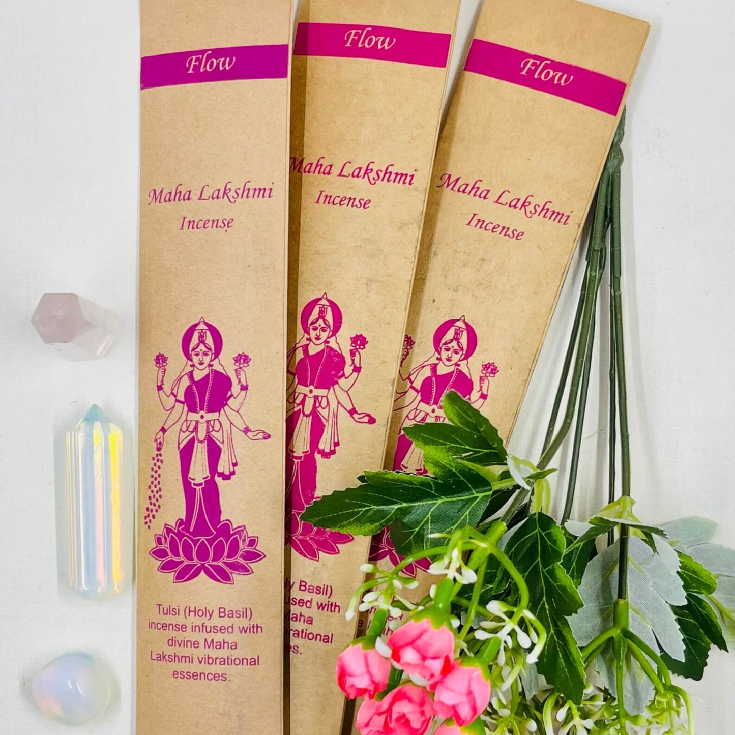 A packet of incense sticks labeled 'Flow' with an illustration of the goddess Maha Lakshmi, accompanied by a small crystal, a candle, and fresh flowers.
