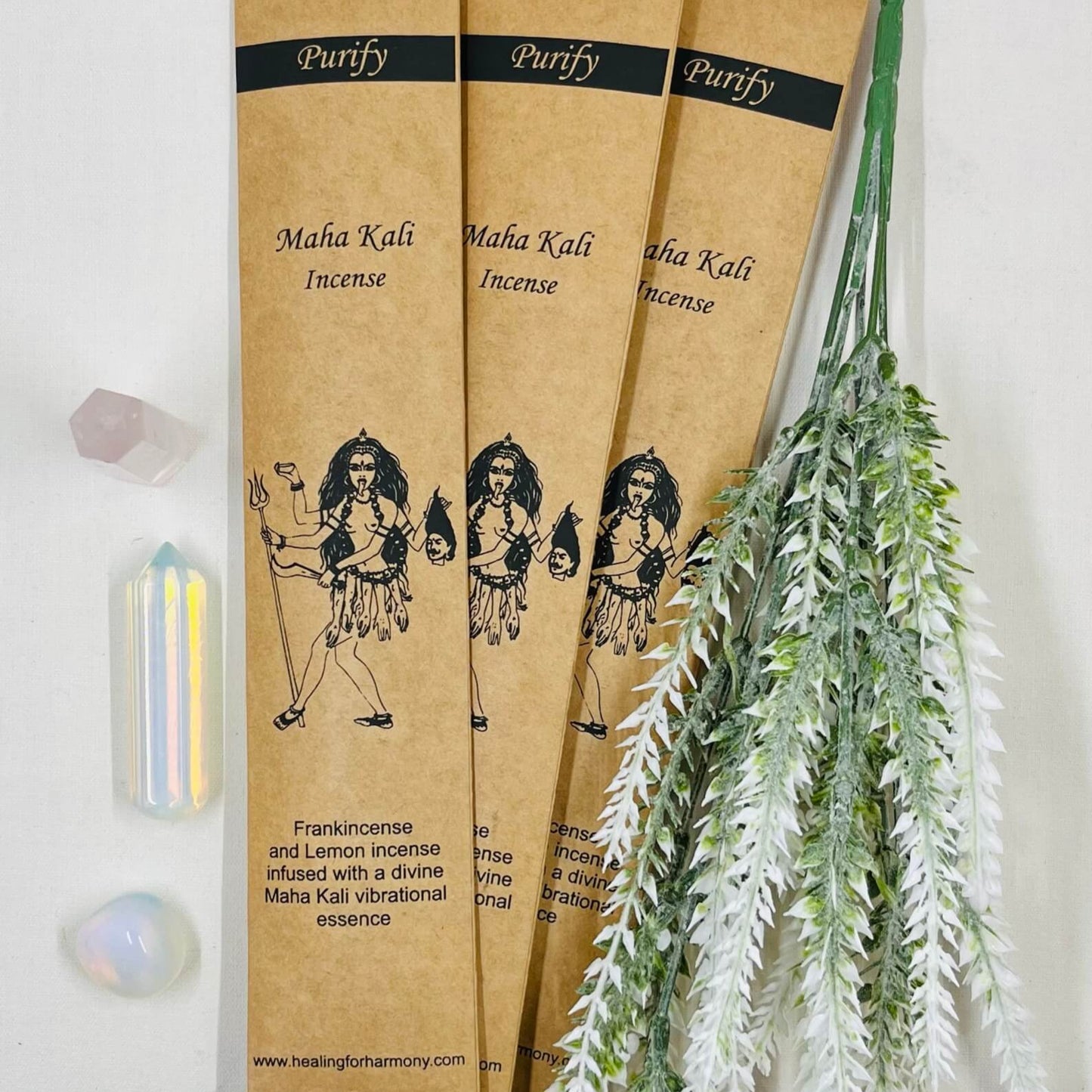 A packet of Maha Kali incense with illustrations, alongside a stick of incense, with decorative herbs and crystals.