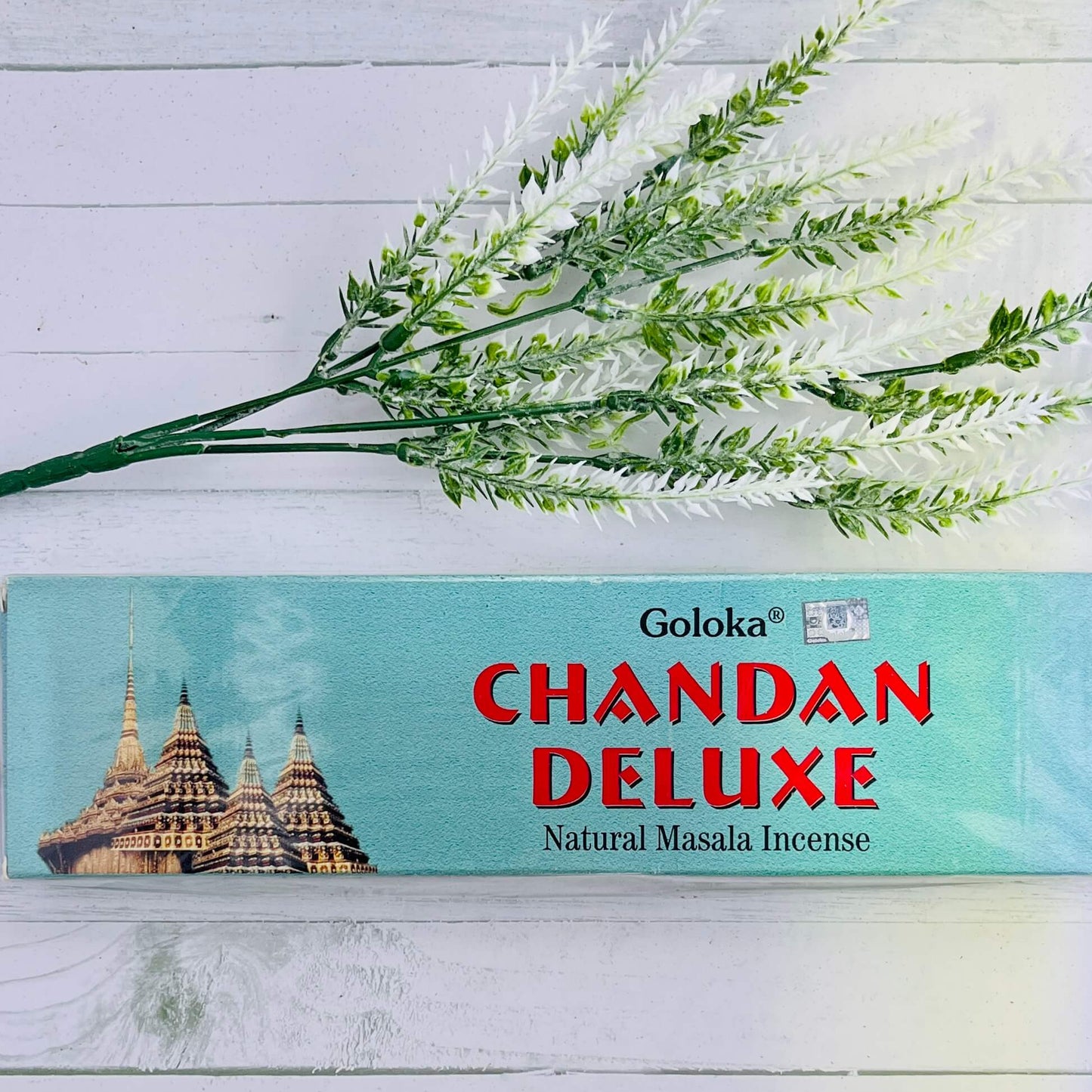 A box of Goloka Chandan Deluxe Natural Masala Incense placed on a white surface with a sprig of dried herbs lying to the side.