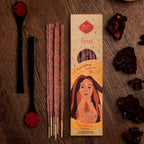 A package of Sagrada Madre incense with several long black sticks and a decorative red dot, alongside a wooden surface with fruit biomass and charcoal pieces for burning the incense.