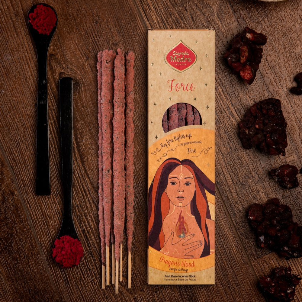 A package of Sagrada Madre incense with several long black sticks and a decorative red dot, alongside a wooden surface with fruit biomass and charcoal pieces for burning the incense.