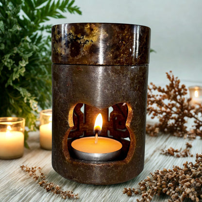 Decorative oil burner with a lit candle on a wooden surface with plants in the background