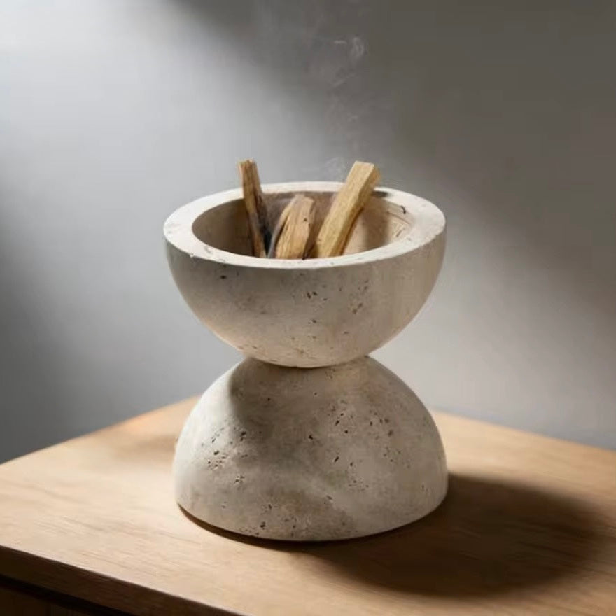 A two-tiered stone incense holder with a textured beige finish, containing several wooden incense sticks, set on a light wood surface against a neutral gray background.
