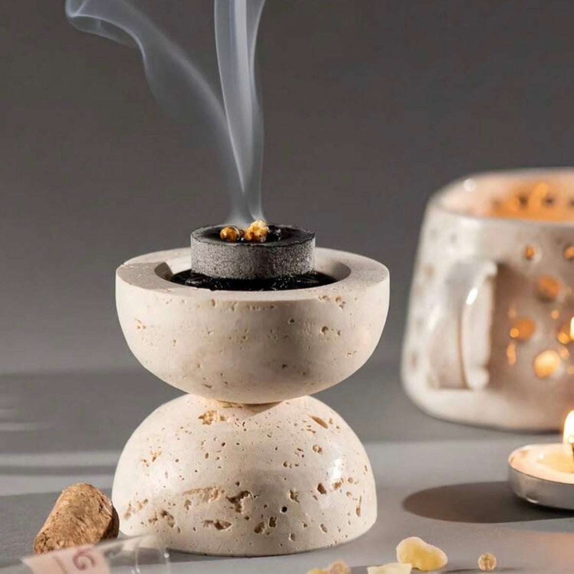 A stone incense burner with a smoking incense holder on top, set on a gray surface with scattered incense pieces and a blurred second burner in the background.