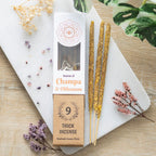A package of Green Tree Champa & Olibanum Botanical Incense Sticks, with 9 incense sticks visible outside the package. The packaging is brown with an illustration of incense sticks and a decorative design around it, placed on a light surface with flowers and a plant in the background.