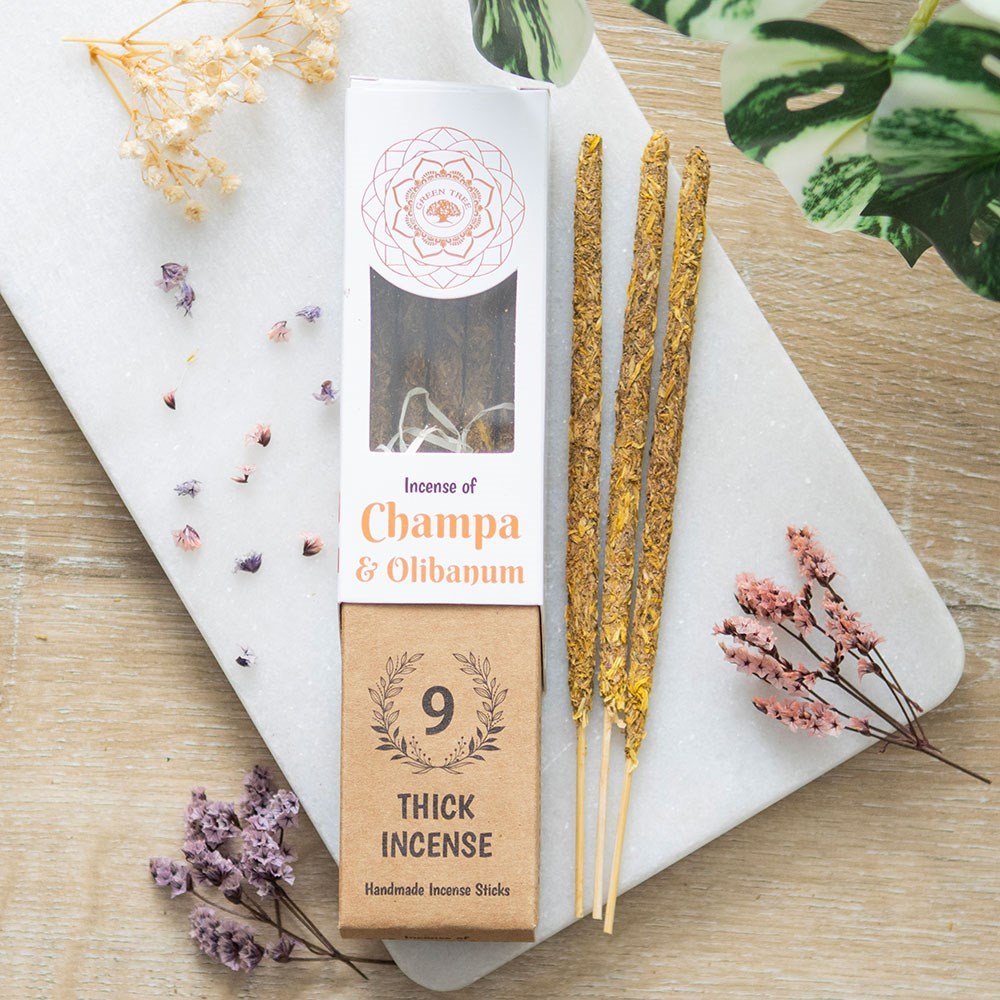 A package of Green Tree Champa & Olibanum Botanical Incense Sticks, with 9 incense sticks visible outside the package. The packaging is brown with an illustration of incense sticks and a decorative design around it, placed on a light surface with flowers and a plant in the background.
