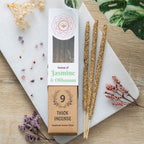 A package of Green Tree Jasmine & Olibanum Botanical Incense with nine sticks visible in front, placed on a wooden surface with decorative flowers and a plant in the background.