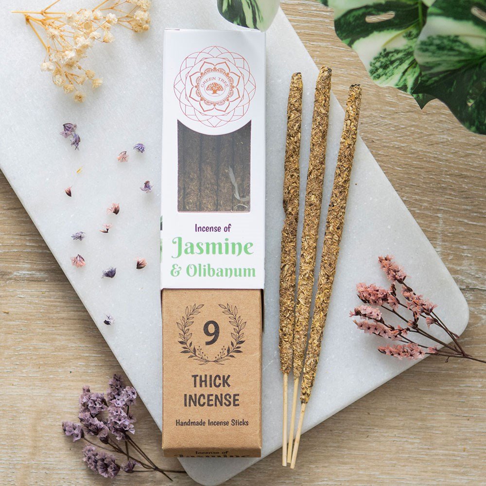 A package of Green Tree Jasmine & Olibanum Botanical Incense with nine sticks visible in front, placed on a wooden surface with decorative flowers and a plant in the background.