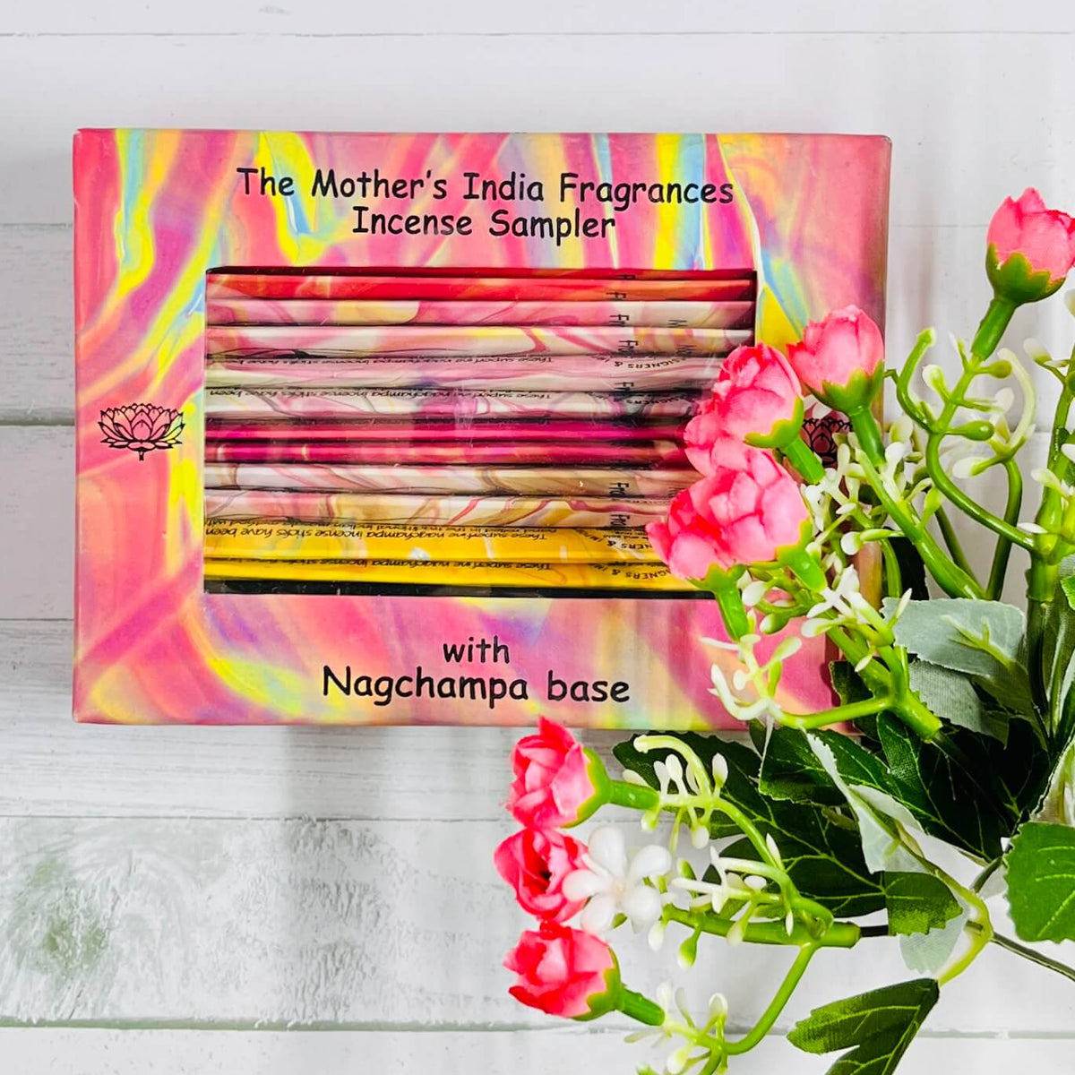 A box of 'The Mother's India Fragrances Incense Sampler' with various incense sticks visible, displayed on a white surface with pink flowers to the side.