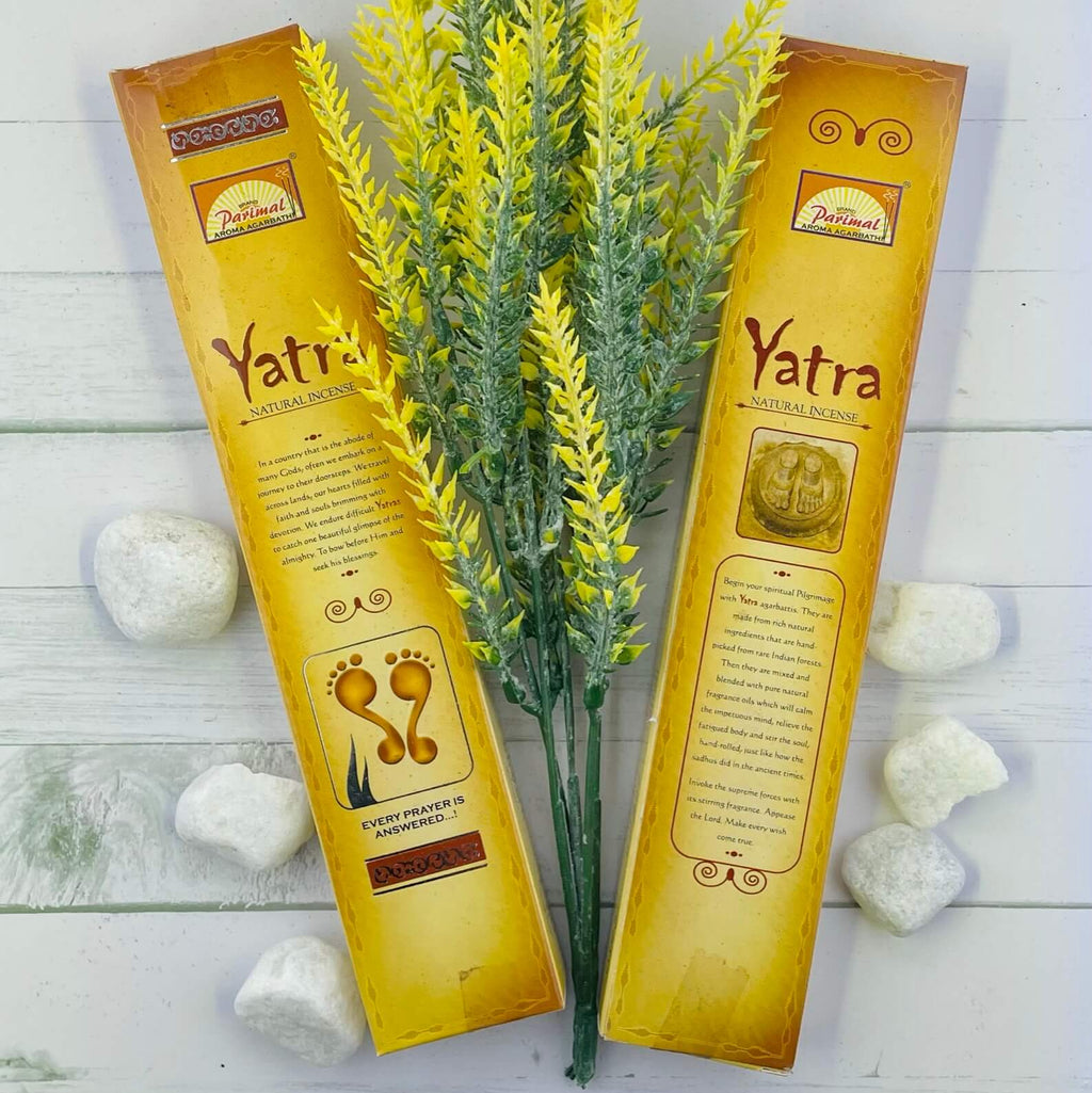 The image shows two packets of Parimal Yatra Natural Incense with a decorative yellow plant and white stones in the background, presented on a white wooden surface.