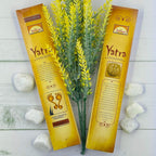 The image shows two packets of Parimal Yatra Natural Incense with a decorative yellow plant and white stones in the background, presented on a white wooden surface.