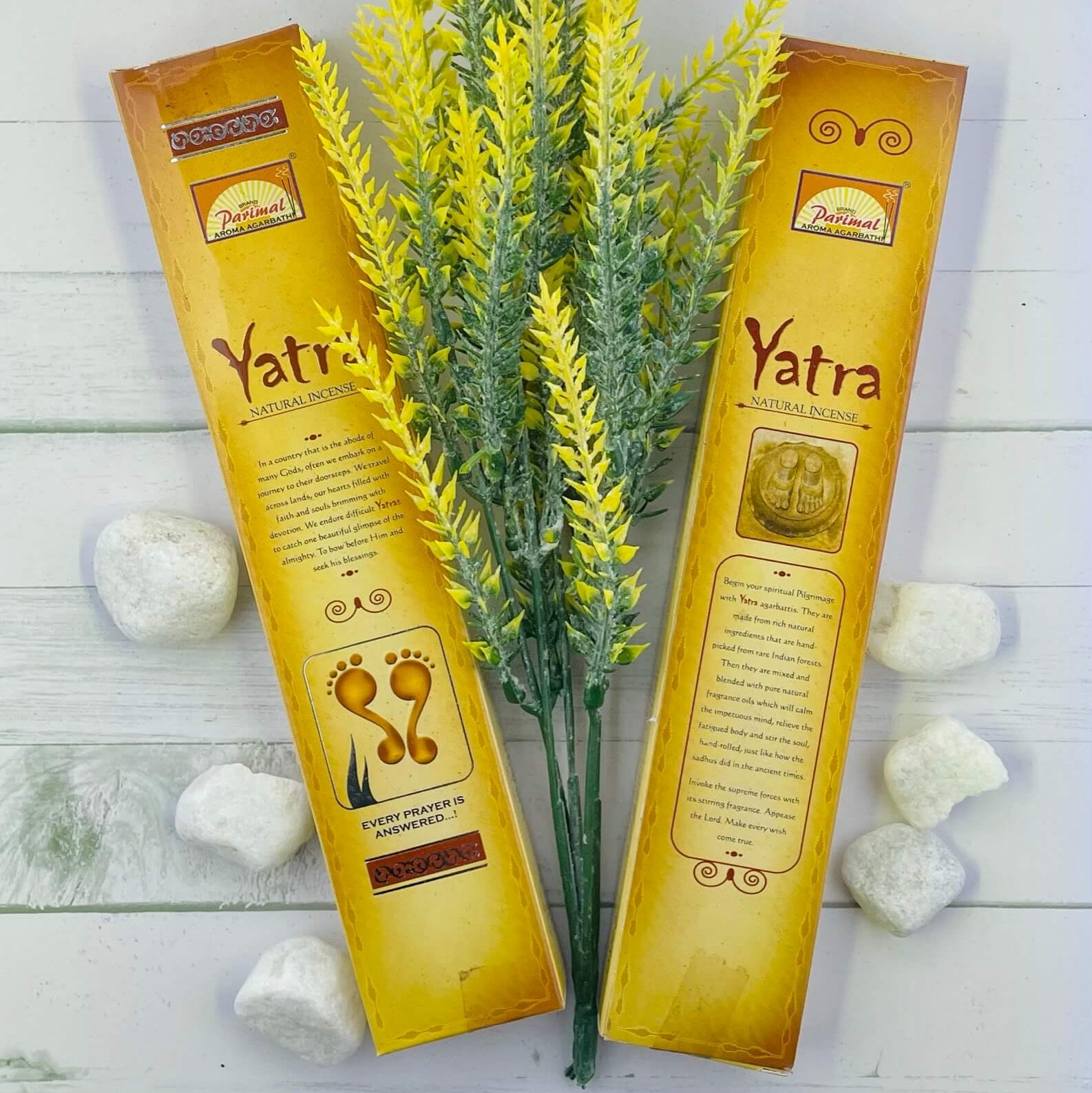 The image shows two packets of Parimal Yatra Natural Incense with a decorative yellow plant and white stones in the background, presented on a white wooden surface.