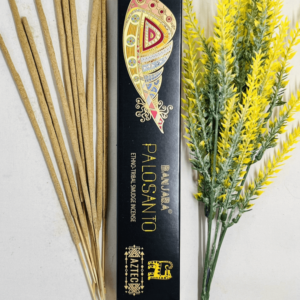 A packet of Banjara Aztec Incense PALO SANTO with several incense sticks displayed in front of it, against a white background with yellow flowers to the side.
