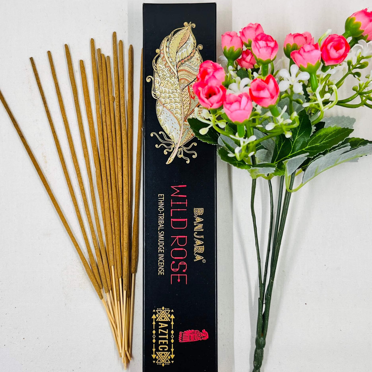 A packet of Banjara Aztec incense sticks with 'WILD ROSE' fragrance, displayed alongside a bouquet of pink roses. The packet is black with gold decorative elements.