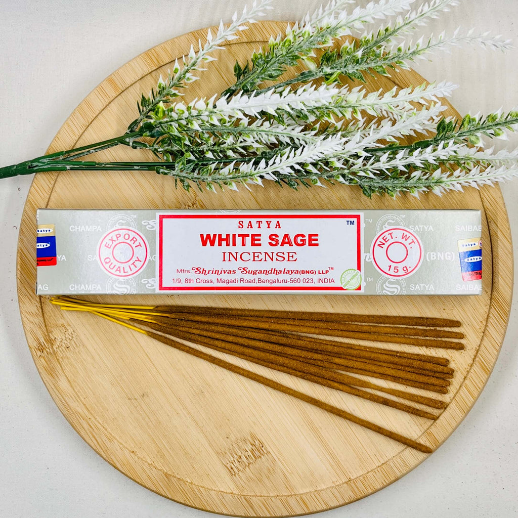 A packet of Satya White Sage incense sticks displayed alongside a sprig of white sage and incense sticks arranged on a wooden surface.