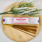 A packet of Satya White Sage incense sticks displayed alongside a sprig of white sage and incense sticks arranged on a wooden surface.