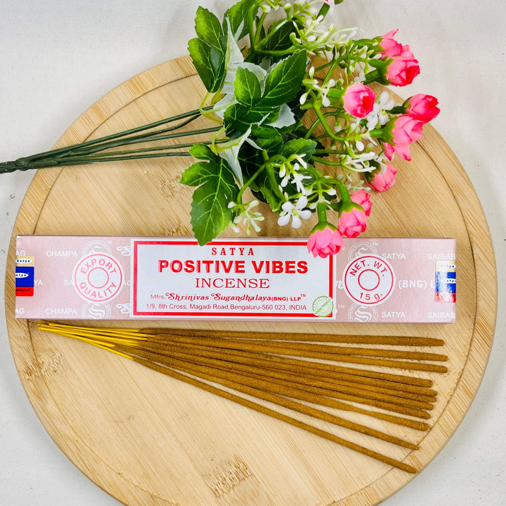 A packet of Satya Positive Vibes incense placed on a wooden surface with a bouquet of pink flowers beside it.