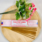 A packet of Satya Positive Vibes incense placed on a wooden surface with a bouquet of pink flowers beside it.