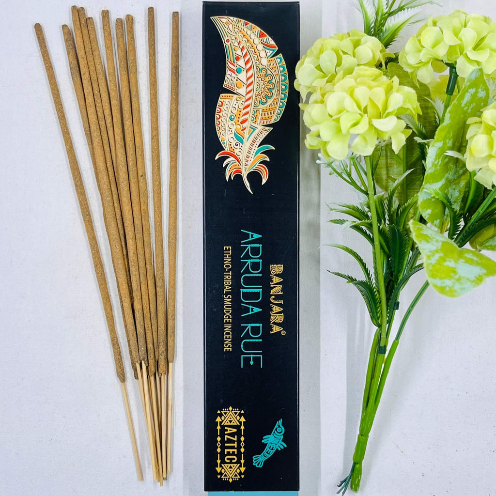 A packet of Banjara Aztec incense sticks with the Arruda Rue fragrance, displayed alongside green artificial flowers. The packaging features a colorful traditional design.