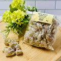A clear plastic package containing medium and large tears of white frankincense resin, with a yellow label, placed on a wooden surface with a bunch of green flowers in the background.