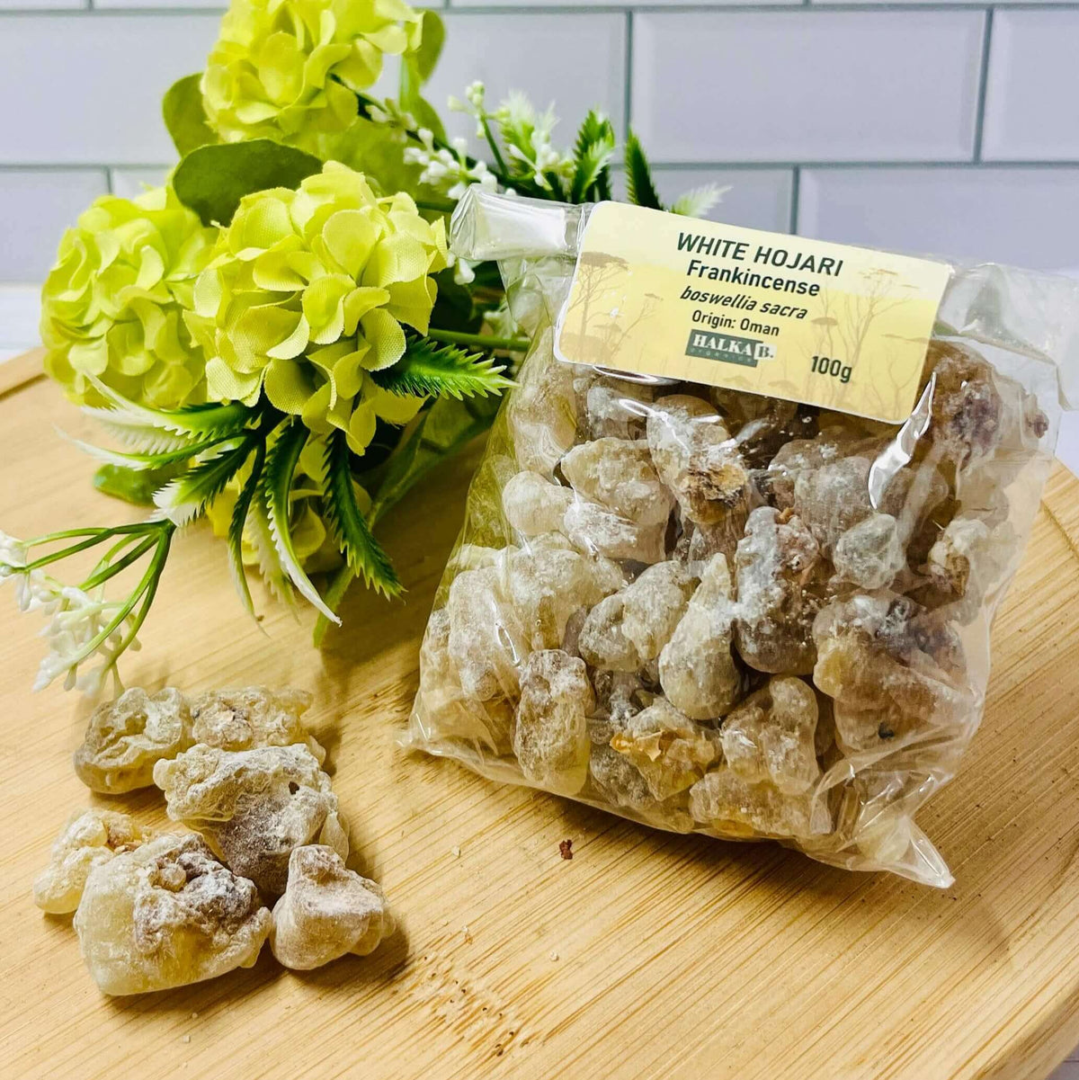 A clear plastic package containing medium and large tears of white frankincense resin, with a yellow label, placed on a wooden surface with a bunch of green flowers in the background.