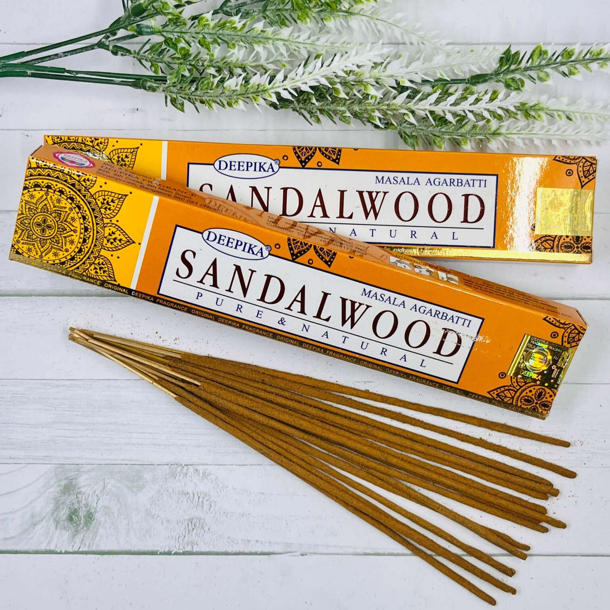 A pack of Deepika Sandalwood incense sticks displayed alongside greenery against a white wooden background.