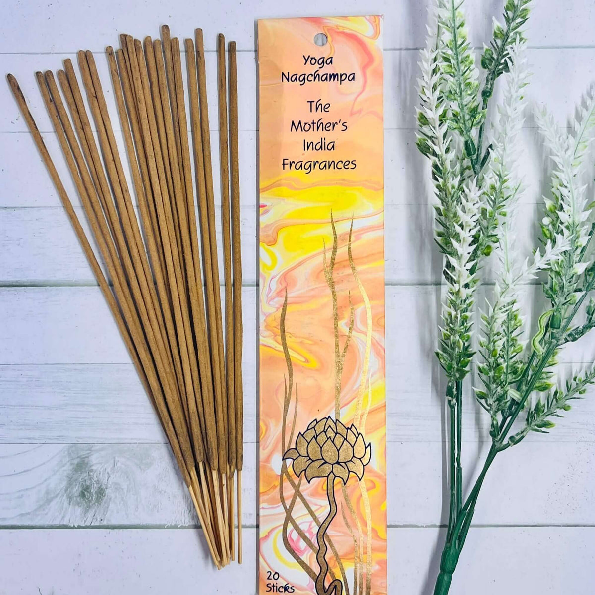A pack of Mother's India Yoga Nag Champa incense sticks displayed alongside a decorative card with a floral and dreamcatcher design, placed on a wooden surface with a sprig of dried herbs to the side.