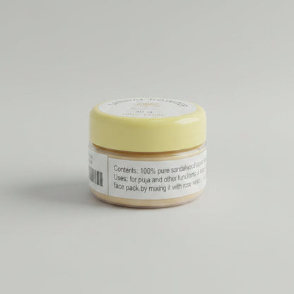 A small round tin of sandal powder sits on a wooden desk with a black keyboard in the background. The lid is pale yellow with a white label reading 'SANDAL POWDER', '30 g', and '100% PURE'.