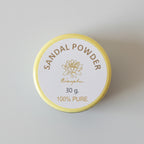 A round tin of Menugha Sandal Powder, 30g, with a yellow rim and a white label, placed on a dark brown wooden surface.