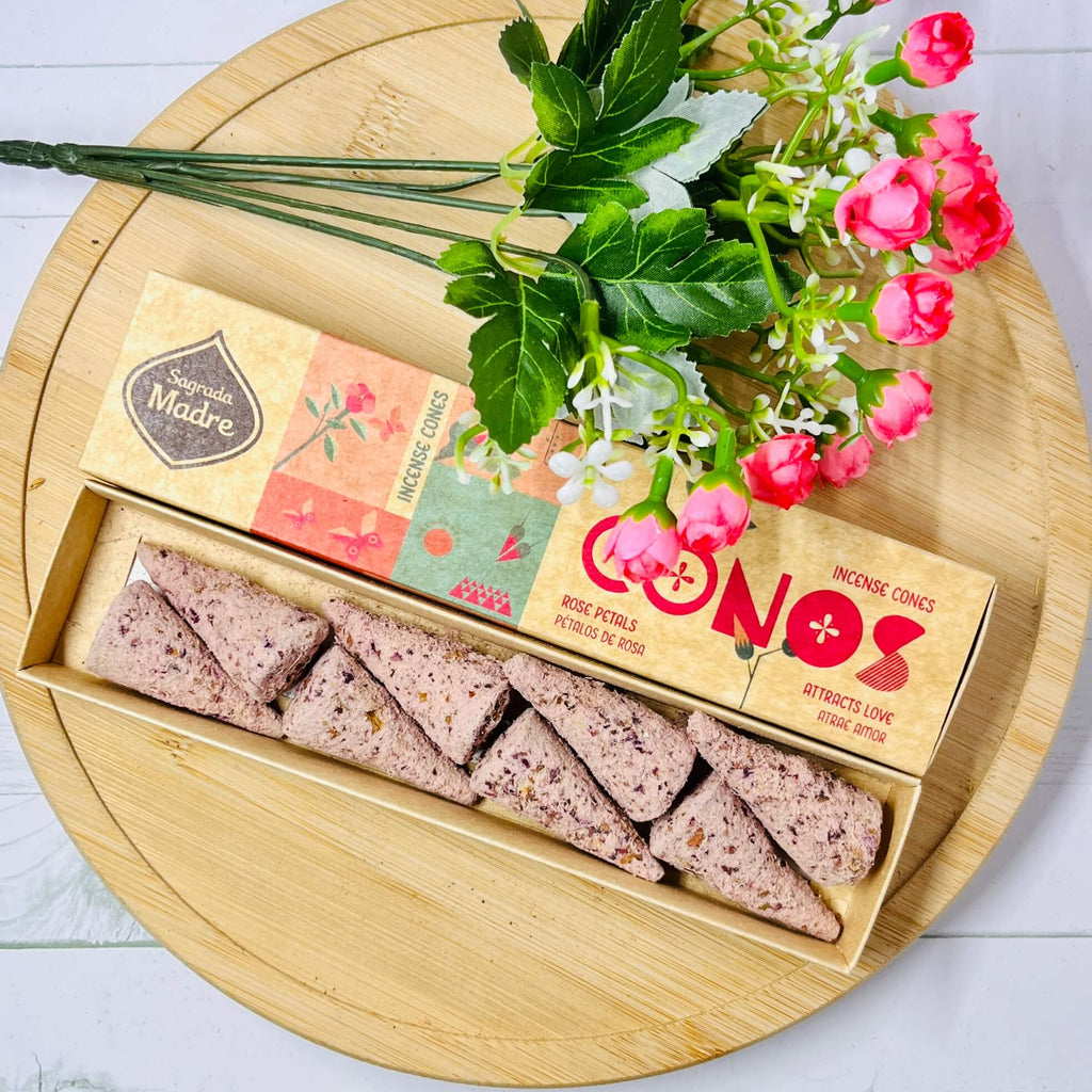 Handmade incense cones from Sagrada Madre displayed next to rose petals and a floral decoration, with the product packaging in the background.