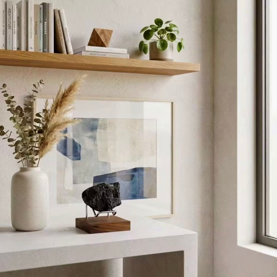 A styled interior corner with a light wood floating shelf holding books and a small potted plant, above a white console table. On the table is a white ceramic vase with dried pampas grass, a framed abstract print, a dark rock specimen on a wooden base, and a window on the right.