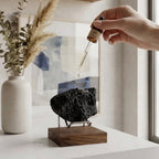 A hand holds a glass dropper above a black porous stone on a wooden stand, with a white vase of dried grass in the background near a window.