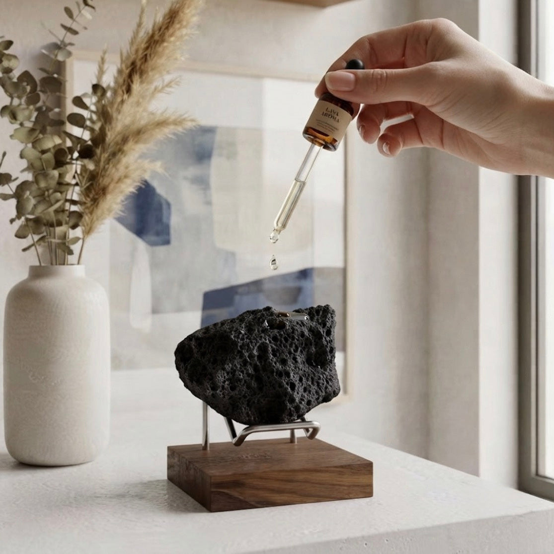 A hand holds a glass dropper above a black porous stone on a wooden stand, with a white vase of dried grass in the background near a window.