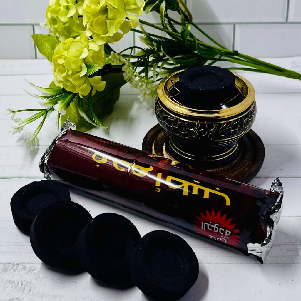 Charcoal discs and incense rolls placed on a white surface with a decorative holder and yellow flowers in the background.
