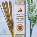 A package of Ayurvedic Palo Santo incense sticks next to a sprig of dried herbs, with the incense stick package displaying the product name and an illustration of a figure meditating.