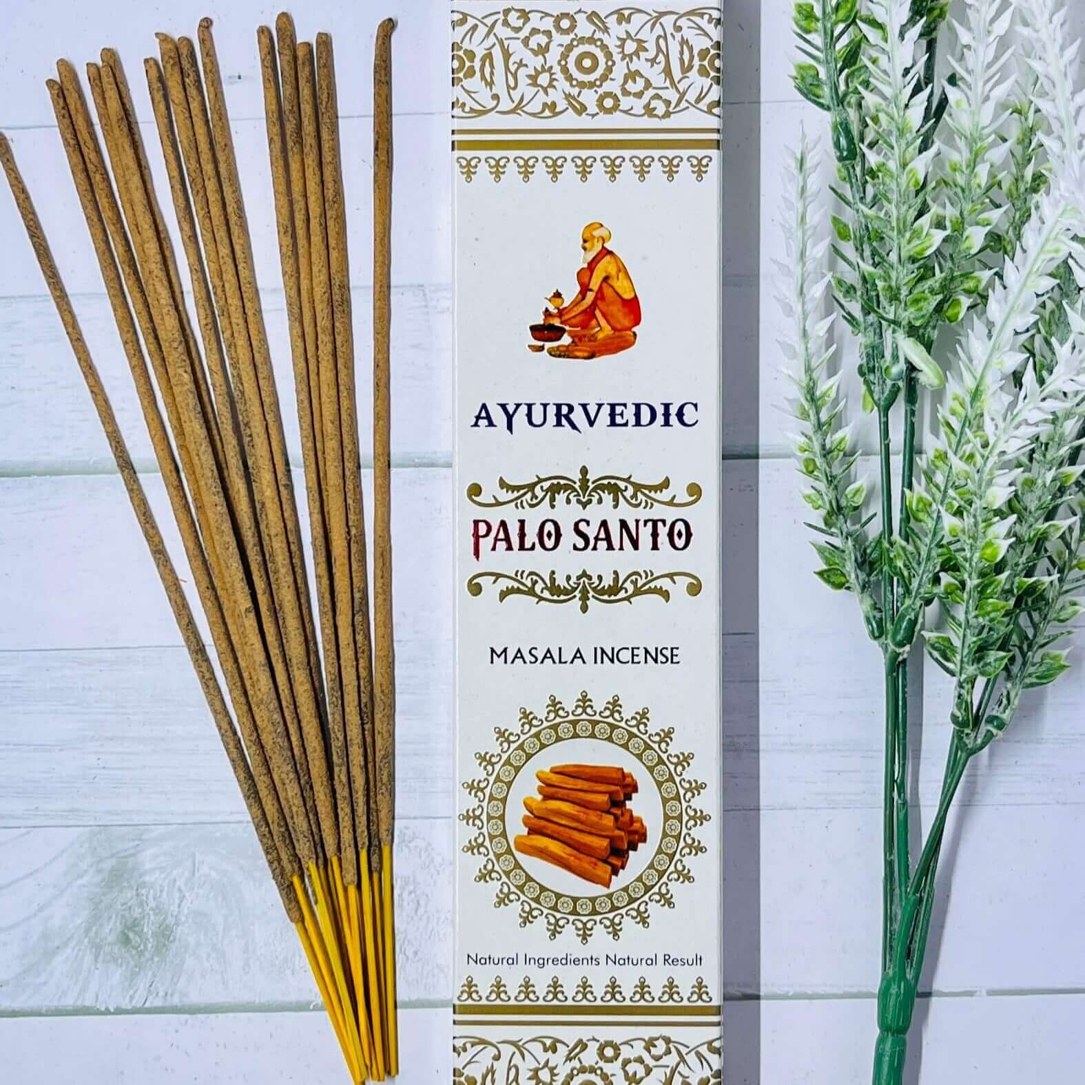 A package of Ayurvedic Palo Santo incense sticks next to a sprig of dried herbs, with the incense stick package displaying the product name and an illustration of a figure meditating.