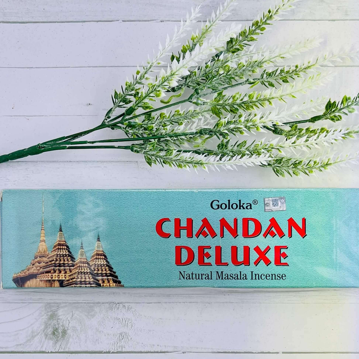 A box of Goloka Chandan Deluxe Natural Masala Incense placed on a white surface with a sprig of dried herbs lying to the side.
