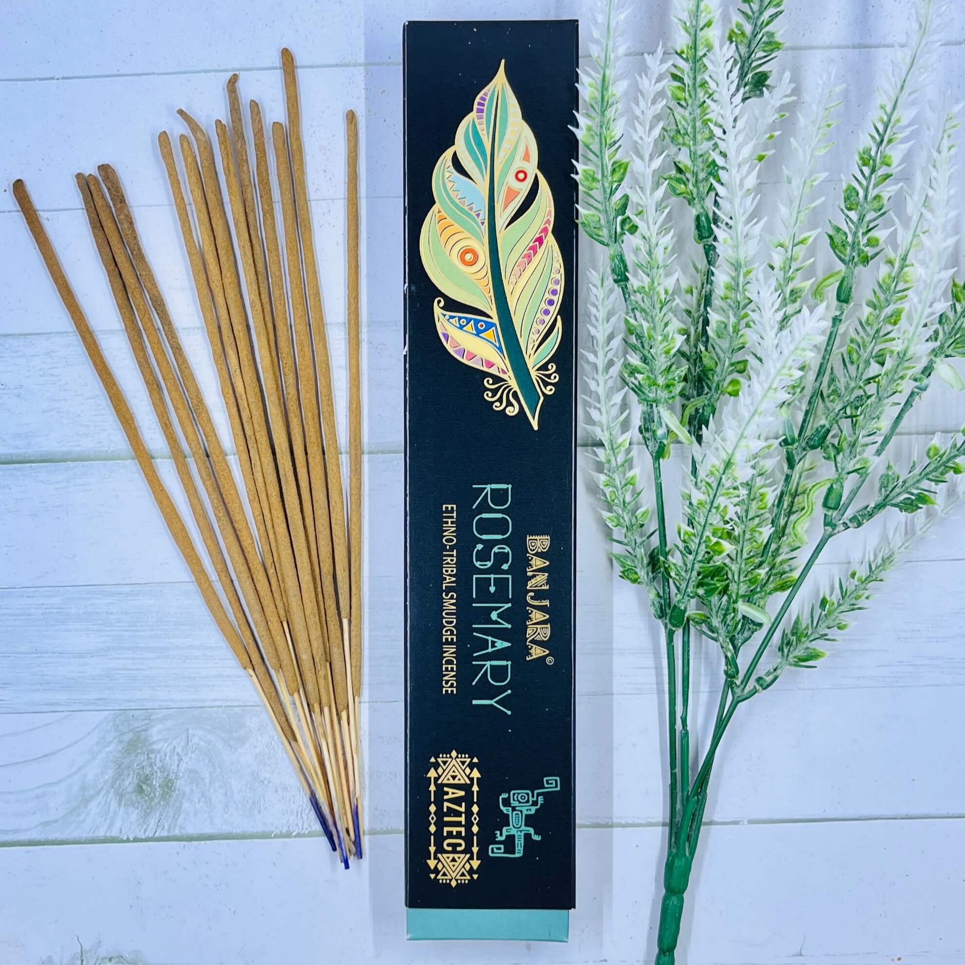 A packet of Banjara Aztec incense sticks with Rosemary fragrance, displayed alongside a decorative feather and rosemary sprig on a white wooden background.
