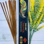 A packet of Banjara Aztec Incense with cinnamon sticks visible in front of the packaging, which is black with orange and white accents and an illustration of a feather. There are yellow flowers to the side.
