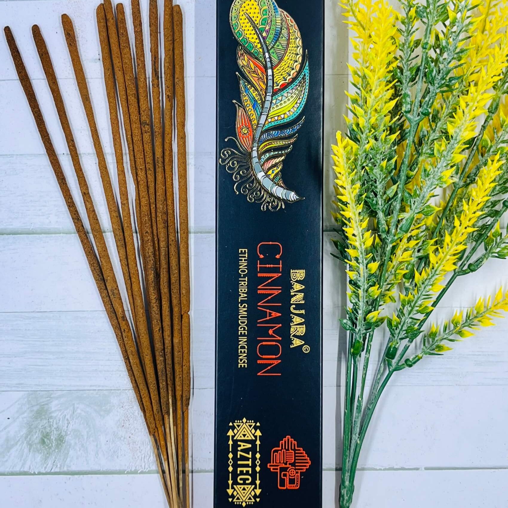 A packet of Banjara Aztec Incense with cinnamon sticks visible in front of the packaging, which is black with orange and white accents and an illustration of a feather. There are yellow flowers to the side.