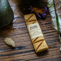 Handmade incense plank on a wooden surface with a label, accompanied by a small piece of natural incense and a pot in the background.