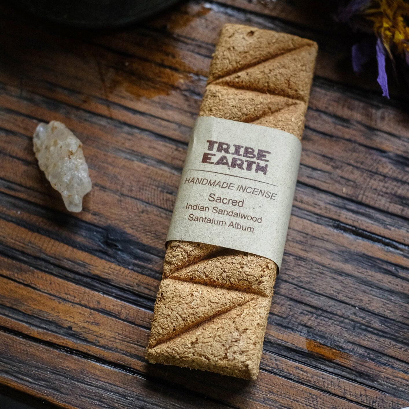 A single plank of handmade Indian Sandalwood incense placed on a wooden surface with a label that reads 'TRIBE EARTH Handmade Indian Sandalwood Incense'. There is also a small crystal next to the incense.
