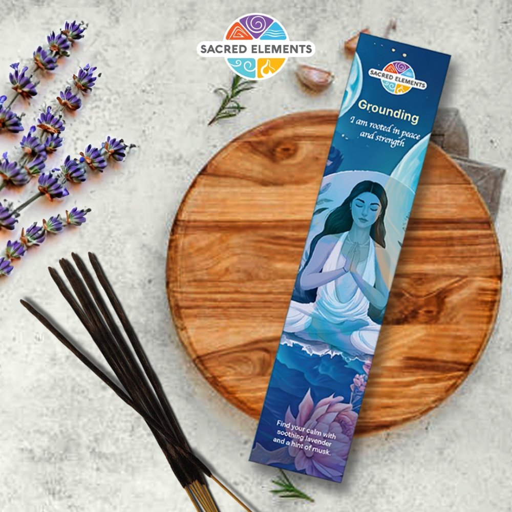 A packet of Sacred Elements GROUNDING Manifestation Incense with an illustration of a woman on it, placed next to some lavender flowers and incense sticks on a wooden board.