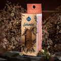 A smudge stick with orange citronella herbs, placed on a rock, with a cardboard packaging in the background that has 'Sagrada Madre' branding and floral designs.