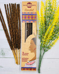 A packet of Sacred Elements Artisan Organic Sandalwood Premium Masala Incense Sticks displayed alongside a bundle of dried flowers. The packet has a colorful design and features an image of a woman's face.