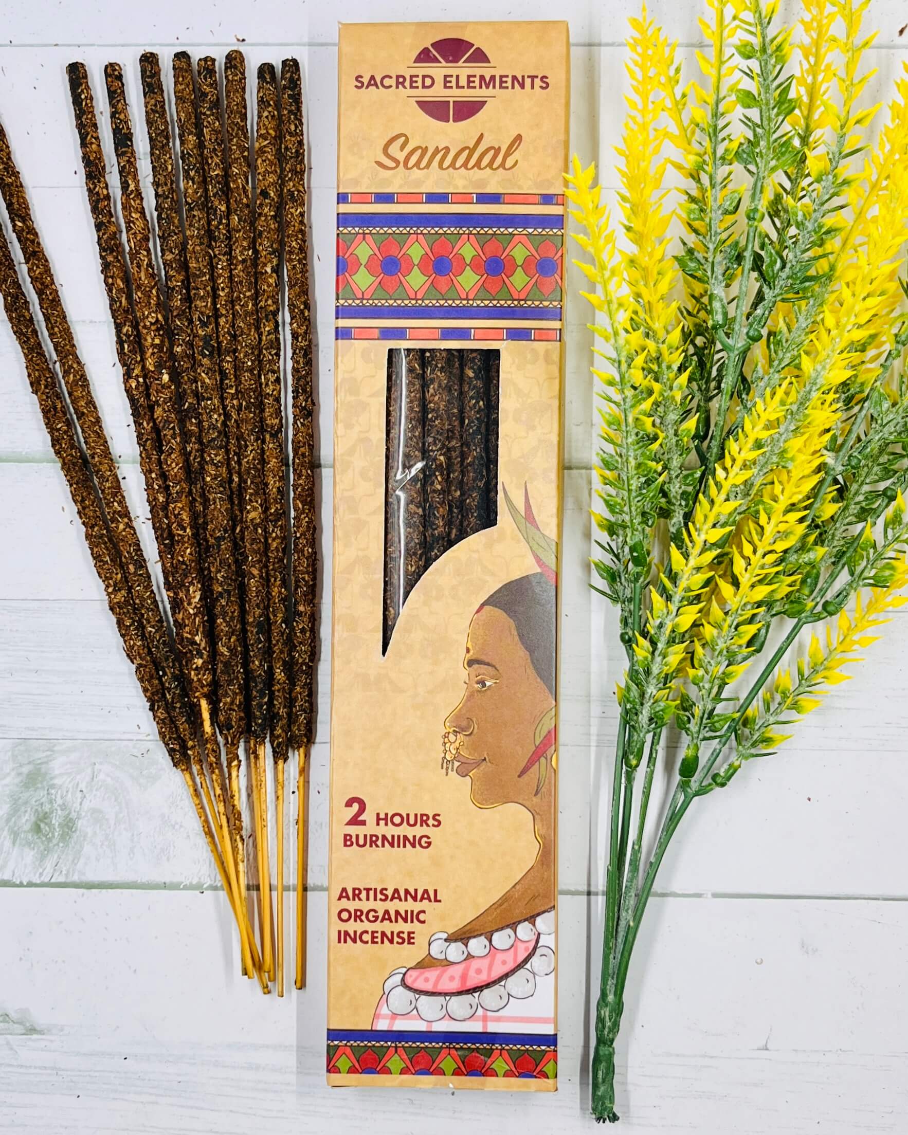 A packet of Sacred Elements Artisan Organic Sandalwood Premium Masala Incense Sticks displayed alongside a bundle of dried flowers. The packet has a colorful design and features an image of a woman's face.