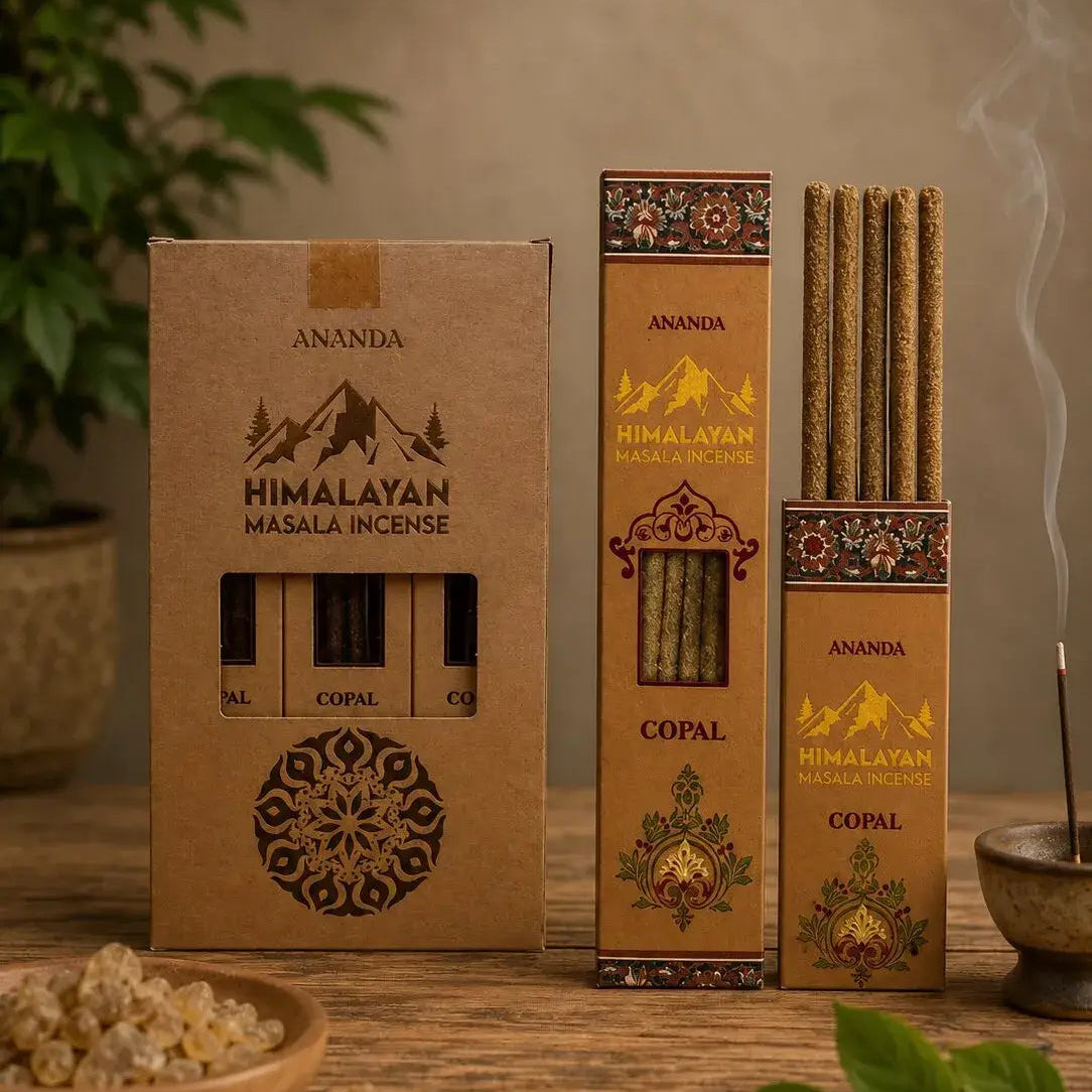 Ananda Himalayan Masala Incense in three brown kraft paper boxes, including one open box showing incense sticks, one box of Copal incense, and several incense sticks standing upright with visible smoke, arranged on a wooden surface with decorative green leaves and a bowl of incense ingredients.