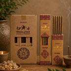 Three boxes of Ananda Himalayan Masala Ingse incense sticks arranged on a wooden surface with a lit candle, incense burner, and green leaves in the background, creating a warm, natural aesthetic.