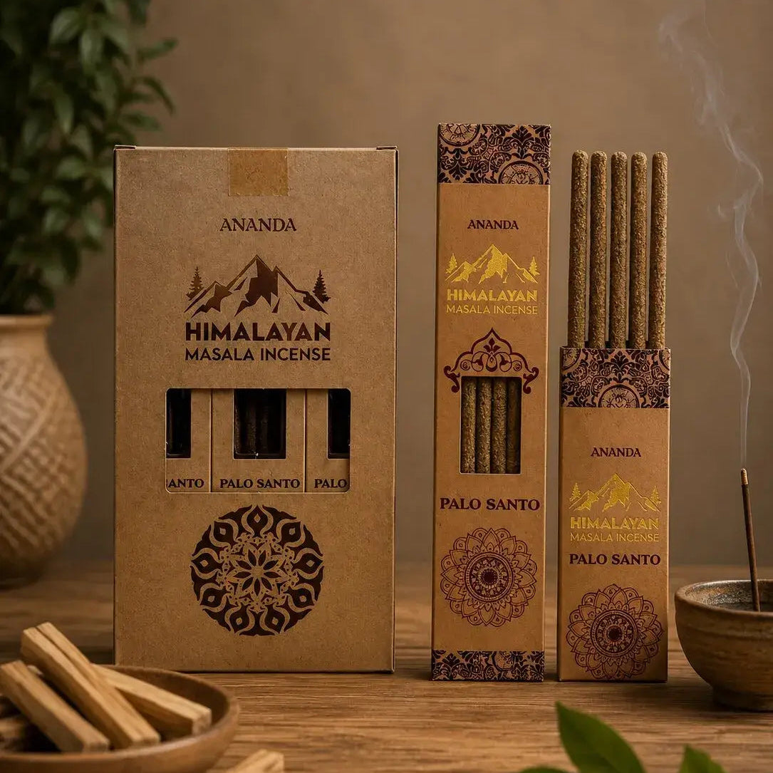 Three boxes of Ananda Himalayan incense in different sizes arranged on a wooden table, with incense sticks visible through the front windows and smoke rising from one box. Decorative leaves, a bowl, and a vase are placed around the products.