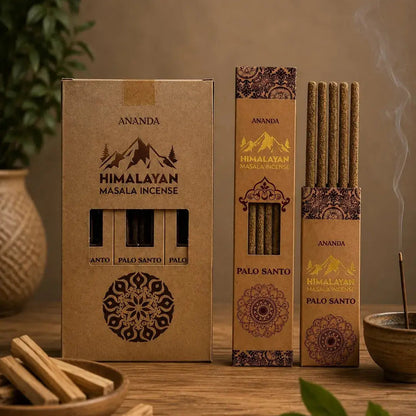 Three boxes of Ananda Himalayan incense in different sizes arranged on a wooden table, with incense sticks visible through the front windows and smoke rising from one box. Decorative leaves, a bowl, and a vase are placed around the products.